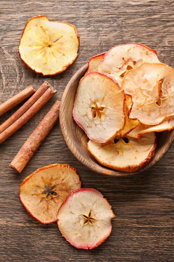 A bowl filled with dried apple slices next to whole dried apple slices and cinnamon sticks on a wooden surface, perfect for those following a comprehensive guide on dehydrating fruits.