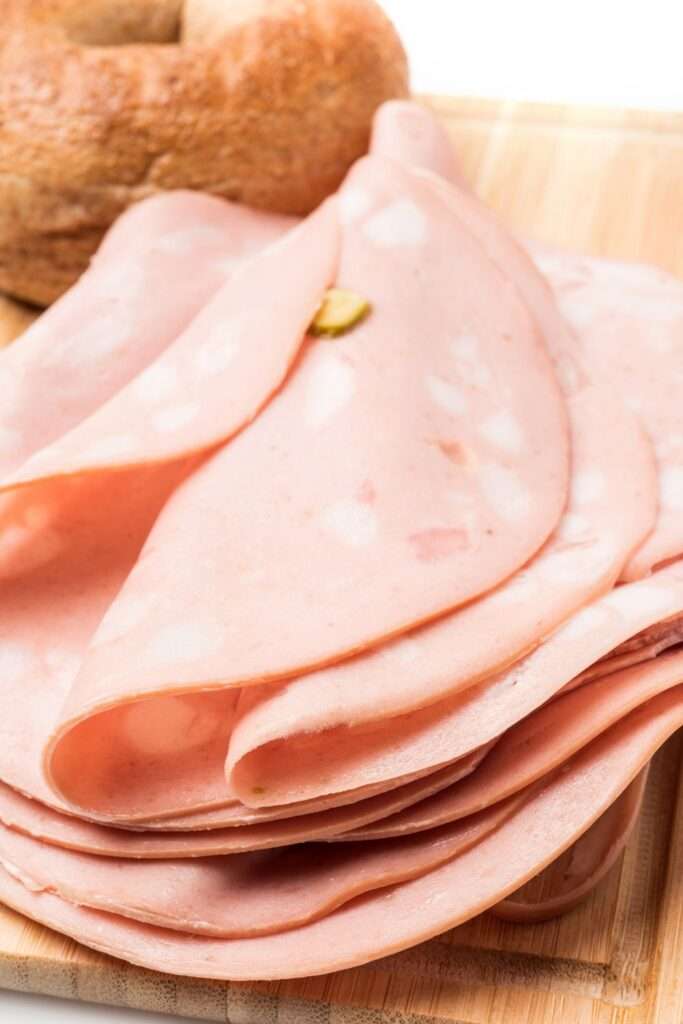 Thin slices of mortadella with a pistachio on a wooden cutting board, accompanied by part of a brown bread loaf, create the perfect charcuterie experience for entertaining.