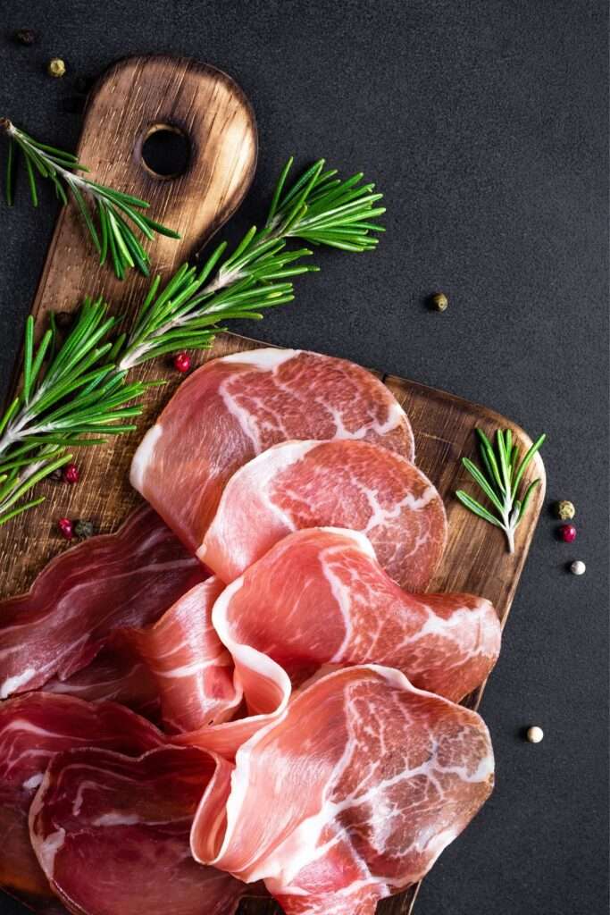 Slices of cured meat arranged on a wooden cutting board with sprigs of rosemary and scattered peppercorns create a delightful charcuterie experience on a dark surface, perfect for entertaining.