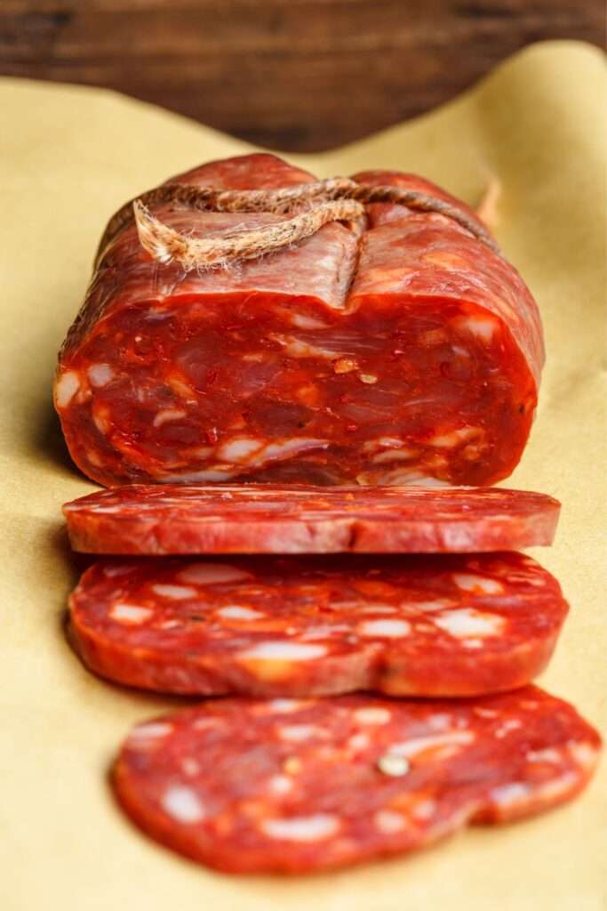 A close-up of a partially sliced cured sausage with a rustic, marbled appearance on a light brown surface, perfect for an elevated charcuterie entertaining experience.