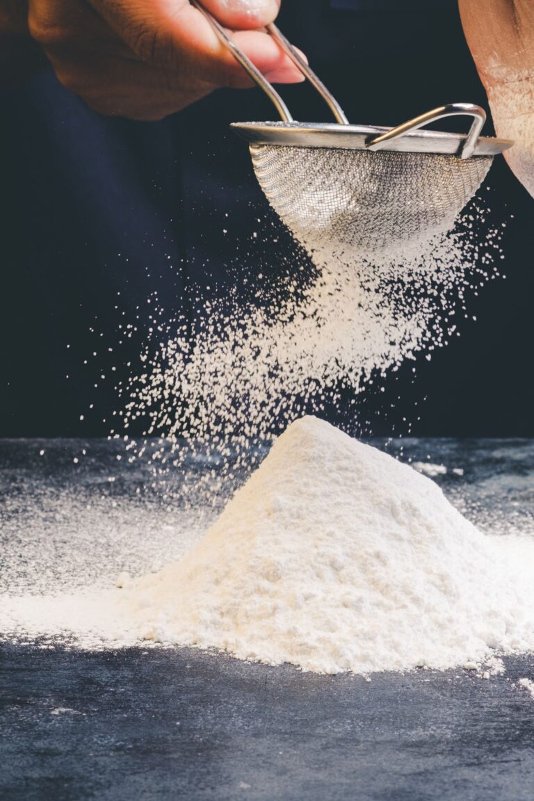A person sifting flour through a fine mesh sieve onto a mound of flour on a dark surface, perfect for creating a Homemade Cake Flour Recipe that results in soft fluffy cakes.